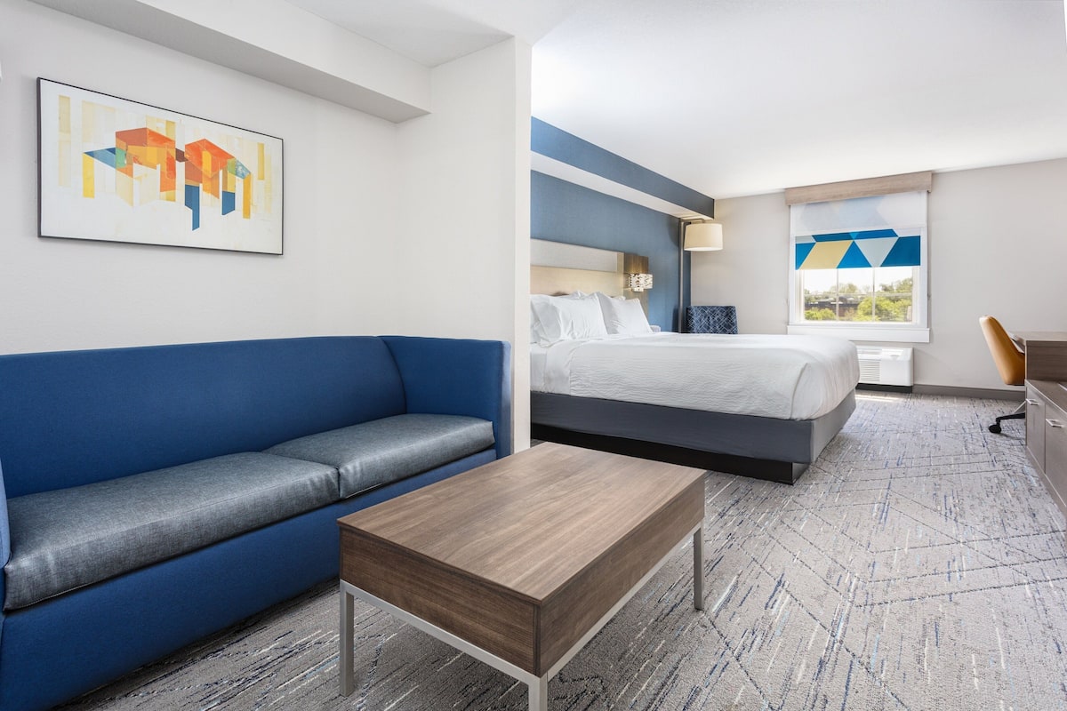 Habitación de hotel con sofá azul, cama con ropa blanca, mesa de centro de madera y un cuadro en la pared.