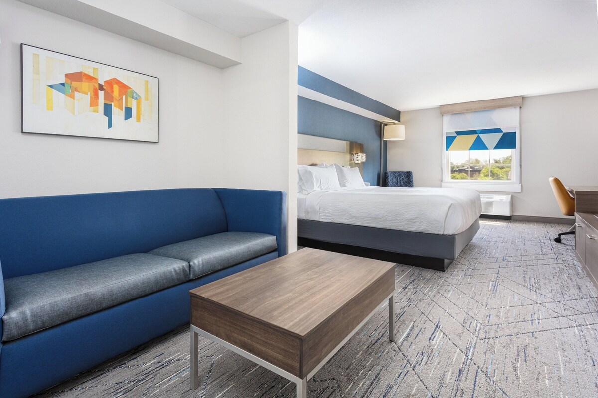 A hotel room with a blue sofa, a bed with white bedding, a wooden coffee table, and a painting on the wall.