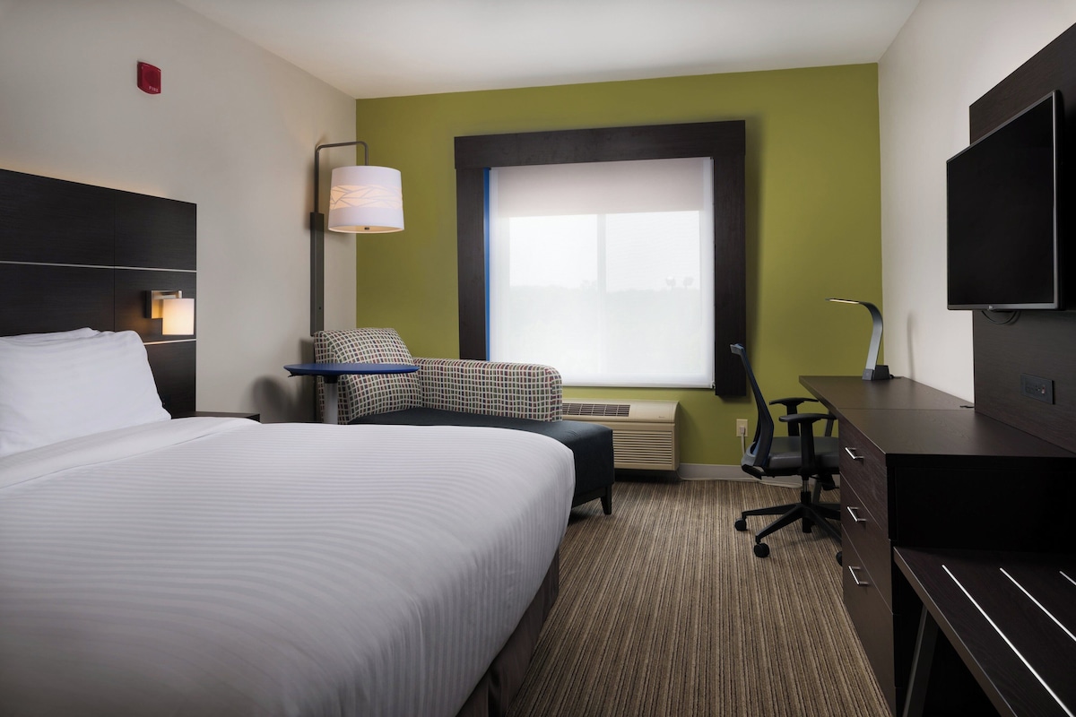 A hotel room with a large bed, a desk, a chair, a television, and a window with blinds.