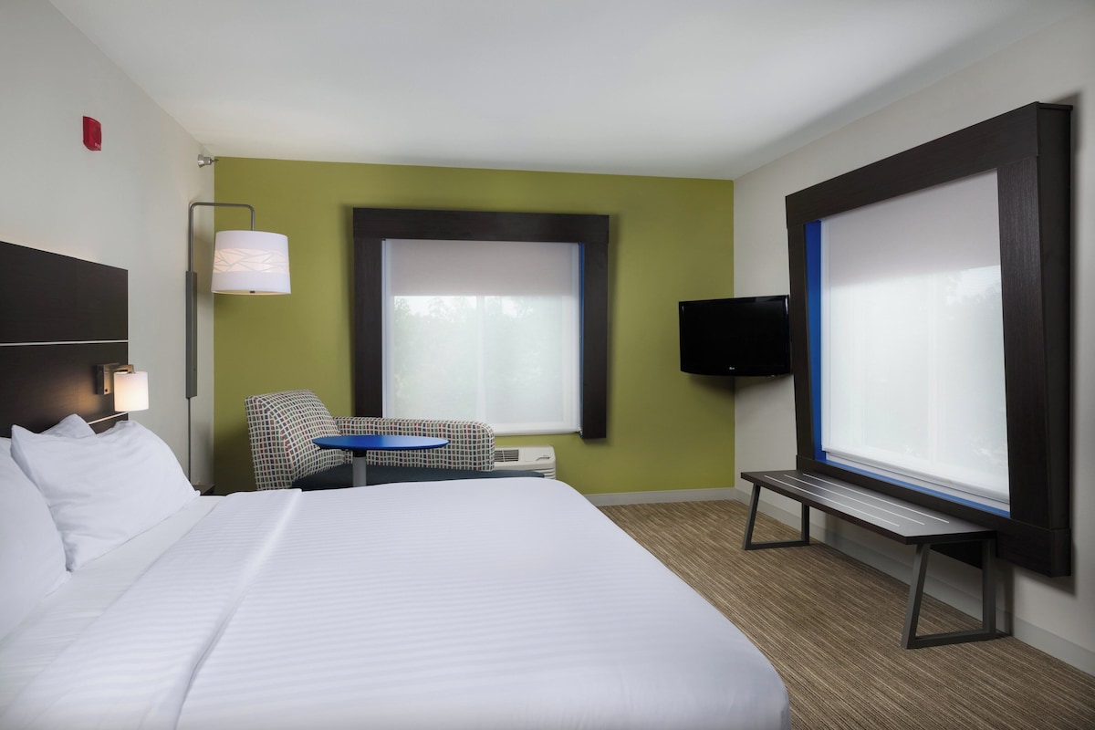 A hotel room with a bed, a chair, a small table, a TV, and a window with blinds.