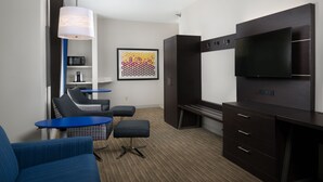 A modern hotel room with a flat-screen TV, dark wood furniture, a blue sofa, a small table, and a colorful artwork on the wall. - Holiday Inn Express Hotel & Suites Independence-Kansas City by IHG (Independence)