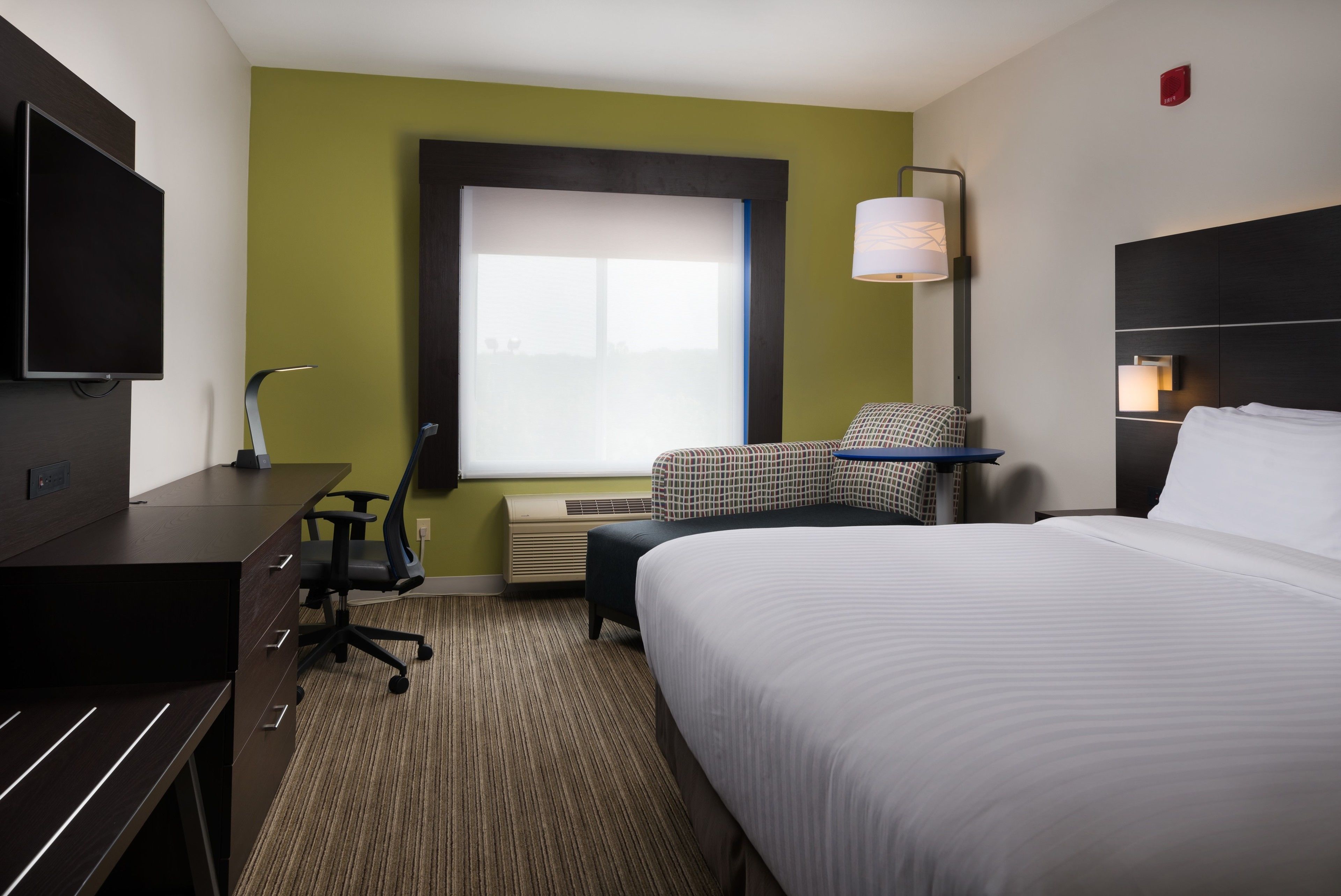 A hotel room with a bed, a desk with a chair, a TV, a window with blinds, and a lamp.
