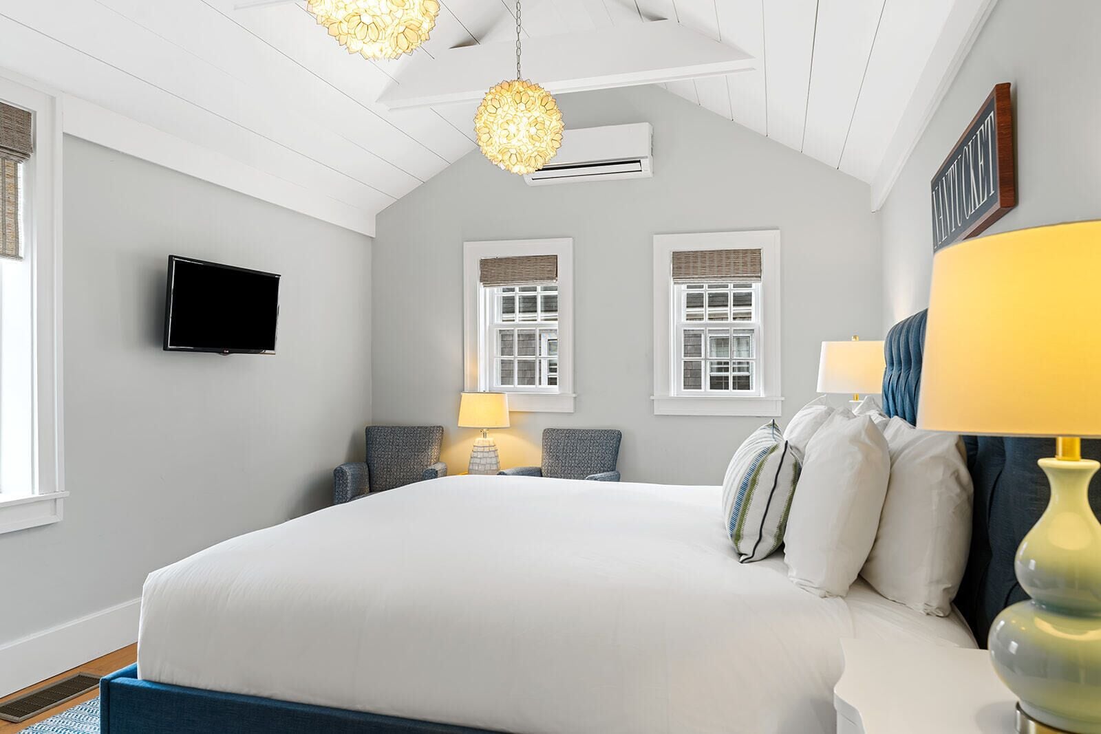 A bedroom with a large bed, two bedside tables with lamps, a flat-screen TV mounted on the wall, and two windows with shutters.