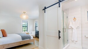 A bedroom with a bed, a bedside table, a hanging light fixture, a sliding door to a bathroom, and a window. - Nantucket Resort Collection (Nantucket)