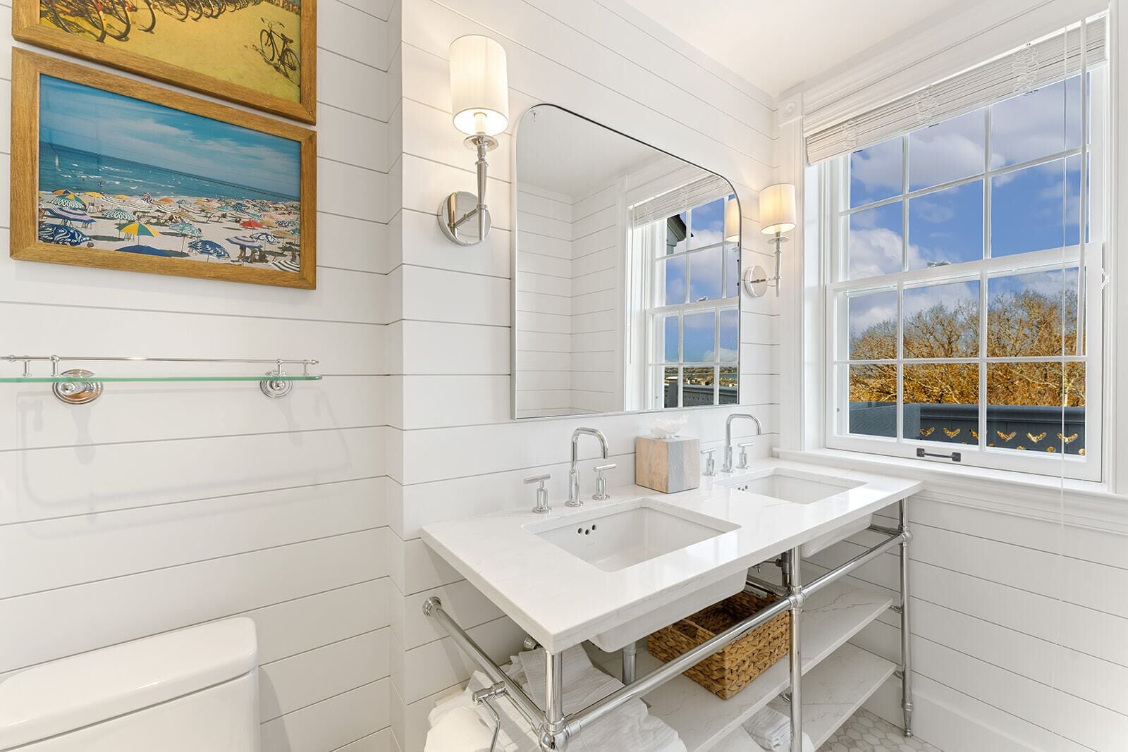 A bathroom with white tiled walls, a double sink, a large mirror, and a window with a view of trees and a building.