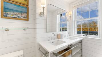 Une salle de bain aux murs recouverts de carreaux blancs, dotée d’un double lavabo, d’un grand miroir et d’une fenêtre donnant sur des arbres et un immeuble.