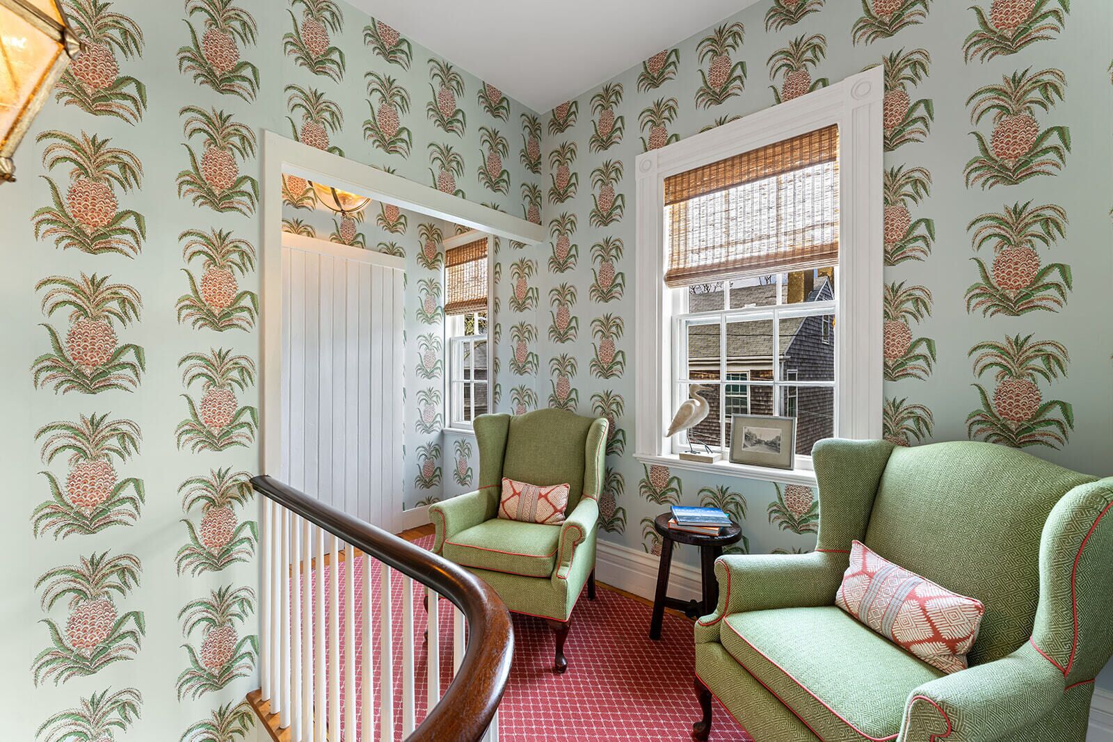 A room with patterned wallpaper, two green armchairs, a wooden staircase, and a window with a view of a house.