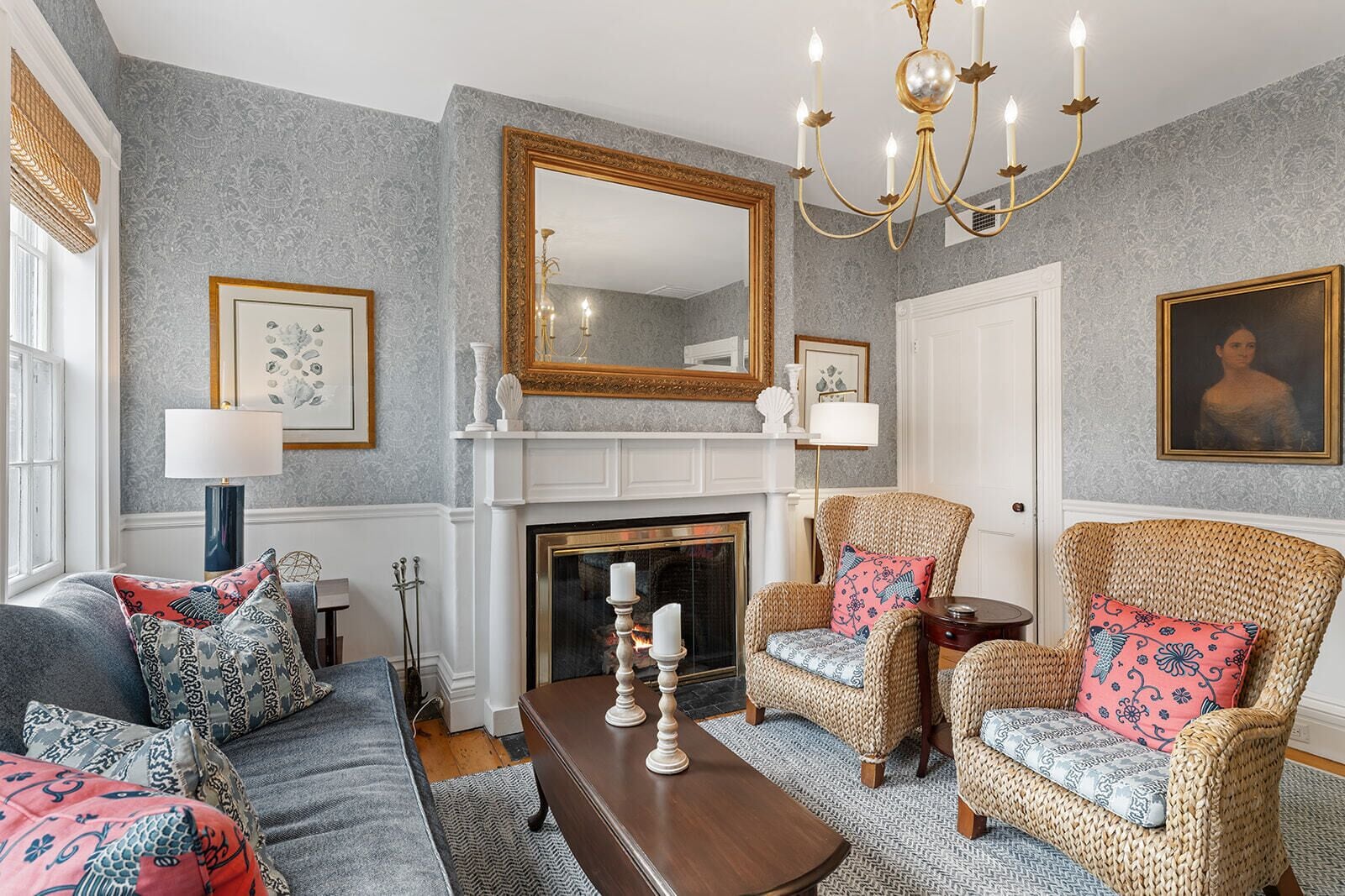 Een woonkamer met een open haard, twee rieten fauteuils met gestreepte kussens, een bank met decoratieve kussens, een houten salontafel en een grote spiegel boven de open haard.