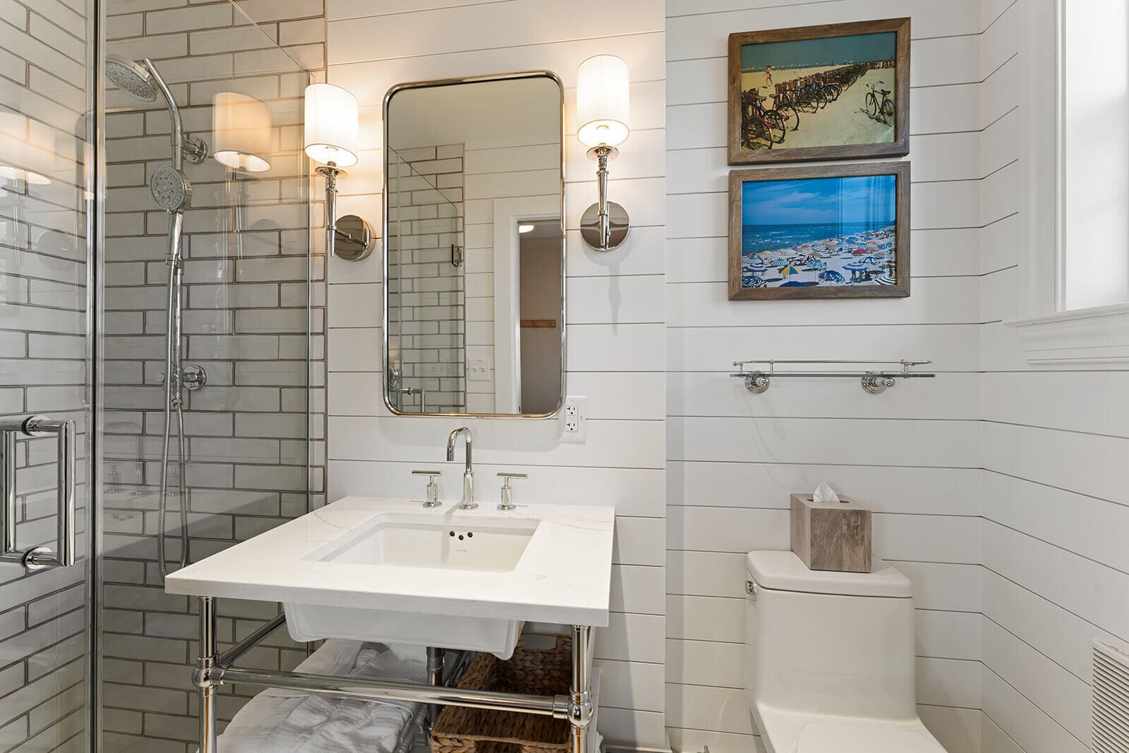 A modern bathroom with a glass-enclosed shower, a white sink with two faucets, and a toilet with a wooden tissue box.
