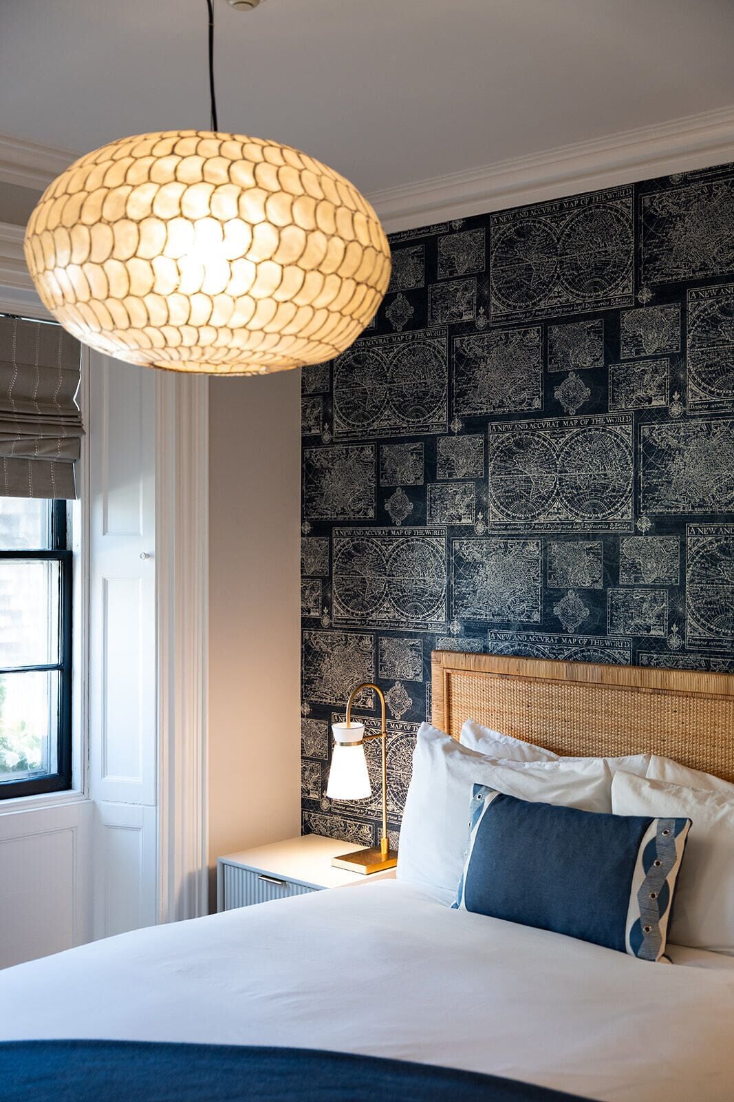 A bedroom with a large bed, a hanging light fixture, a nightstand with a lamp, and a wall with a map pattern.