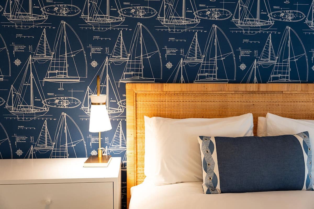 Et hotelværelse med maritime tapeter, en seng med hvide og blå puder og et natbord med en lampe.