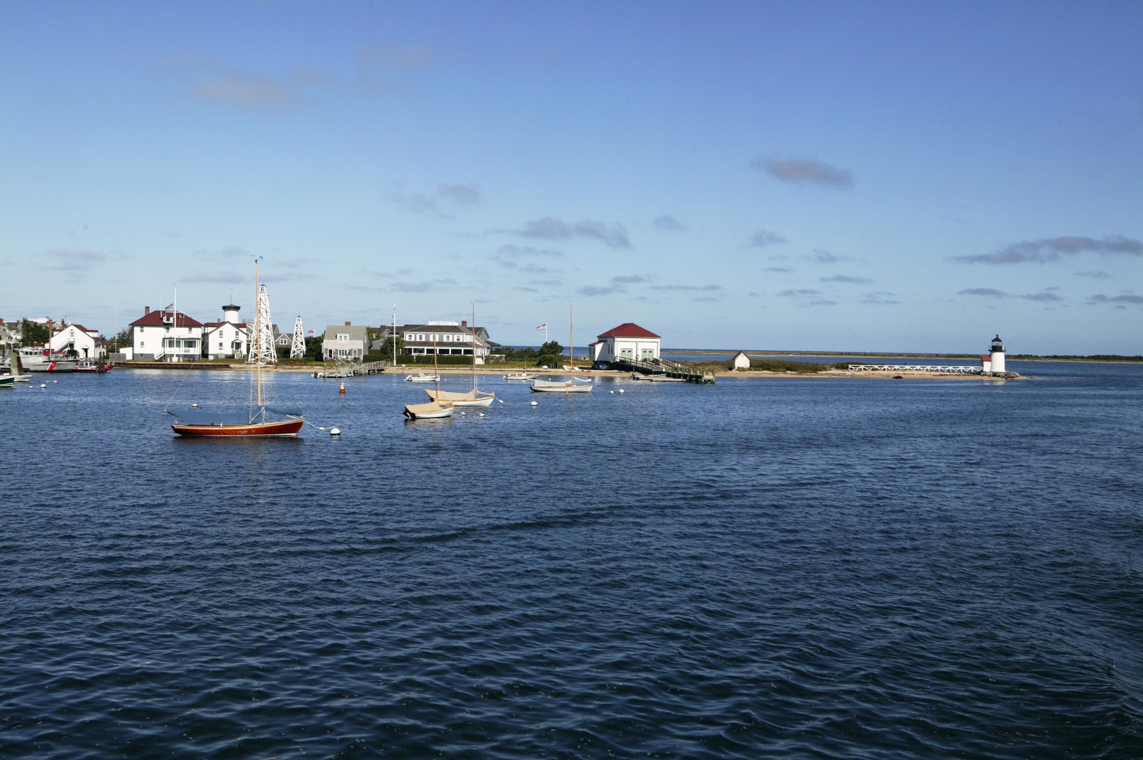 A coastal scene with several boats on calm water, a lighthouse, and buildings along the shore.