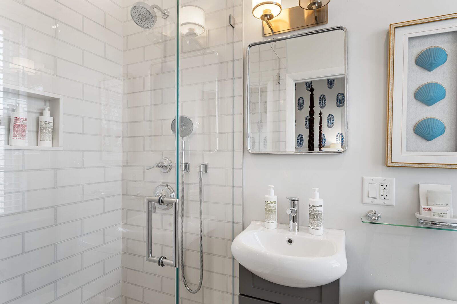 Een moderne badkamer met een douche met glazen wand, een wit wastafelmeubel met twee flesjes MALIN+GOETZ, en een lijst met foto van schelpen aan de muur.