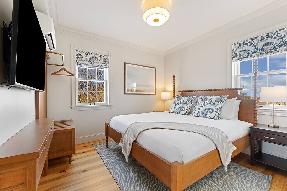 Et pænt opredt værelse med en træseng, et fjernsyn, et skrivebord og et vindue med mønstrede gardiner.