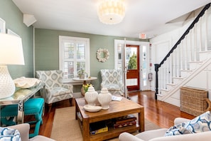 A cozy living room with a wooden coffee table, patterned armchairs, a teal ottoman, and a staircase leading to a second floor.