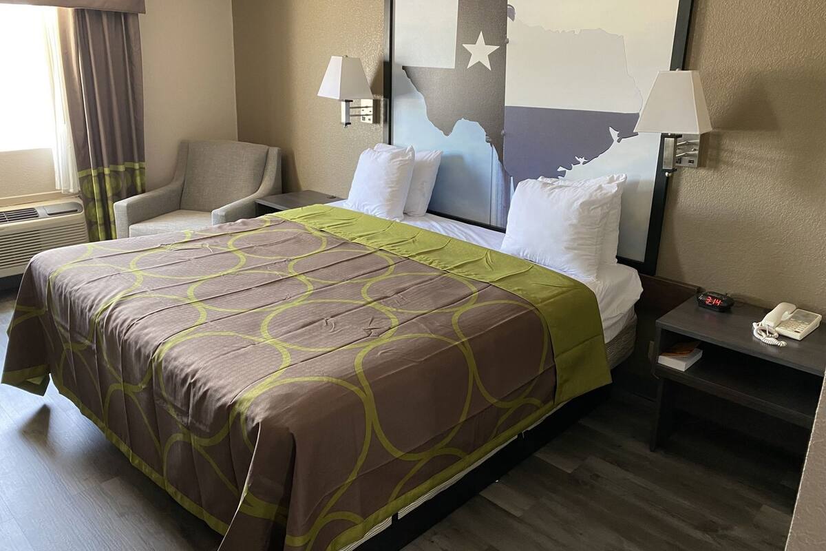 Ein Hotelzimmer mit einem Bett, zwei Nachttischlampen, einem Sessel, einem Nachttisch mit Telefon und Buch sowie einer Wand mit einer großen Landesflagge.