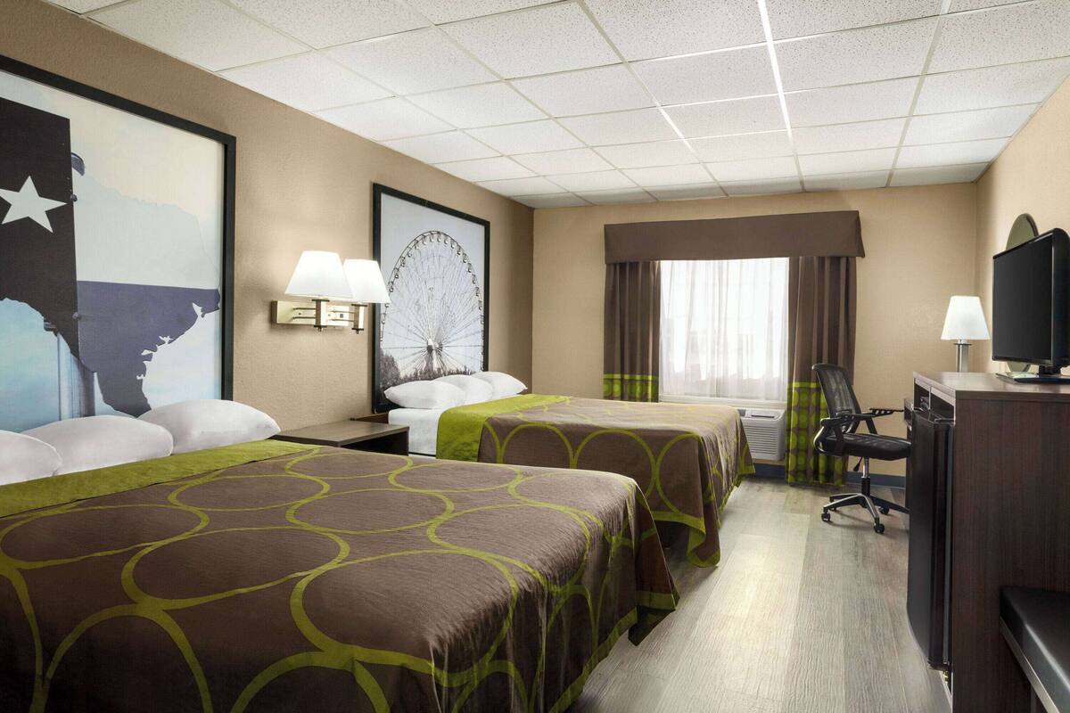 Ein Hotelzimmer mit zwei Betten, einem Schreibtisch, einem Stuhl, einem Fernseher und einem großen Fenster mit Vorhängen.