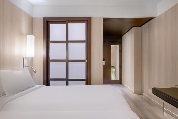 A hotel room with a bed, a bedside lamp, a wardrobe, and a door leading to another room.