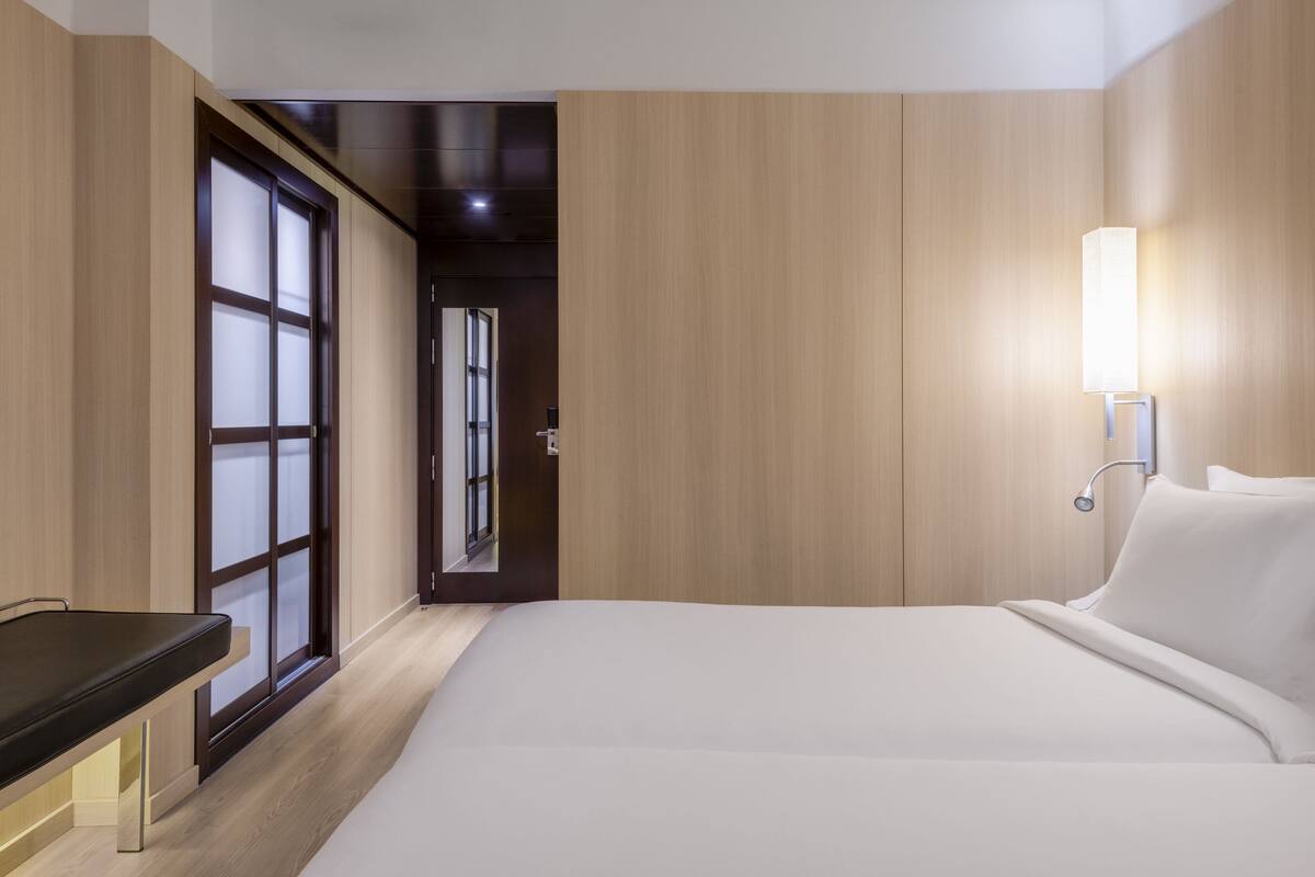 A hotel room with a bed, bedside table, and a wall-mounted lamp.