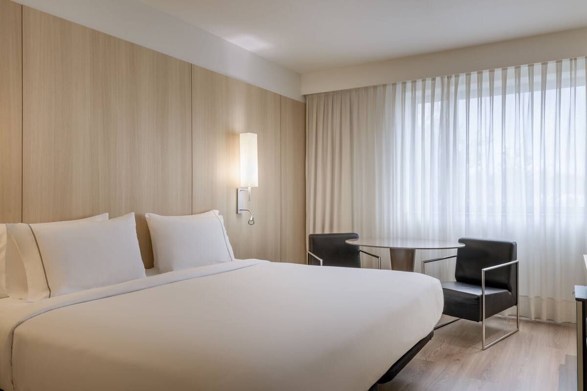 A hotel room with a large bed, a desk, a chair, and a window with curtains.