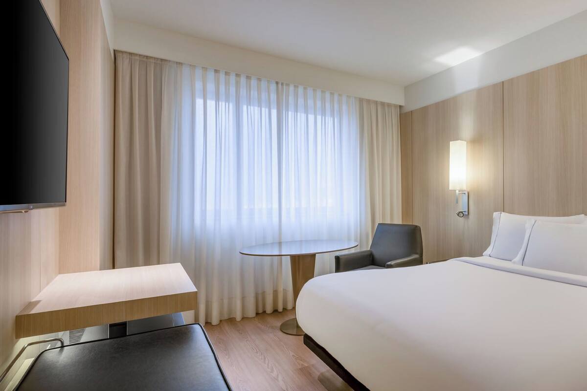 A hotel room with a large bed, a desk, a chair, a TV, and a window with curtains.