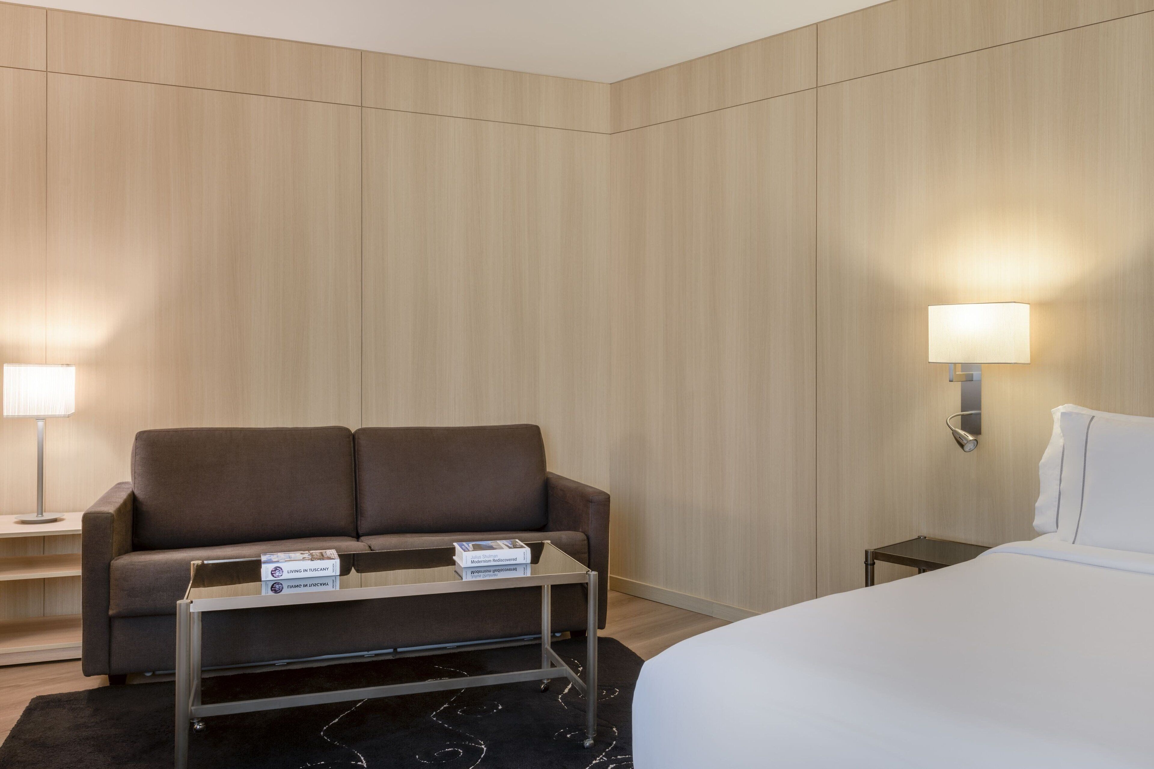 Una habitación de hotel con una cama, un sofá, una mesita y dos lámparas fijadas a la pared.