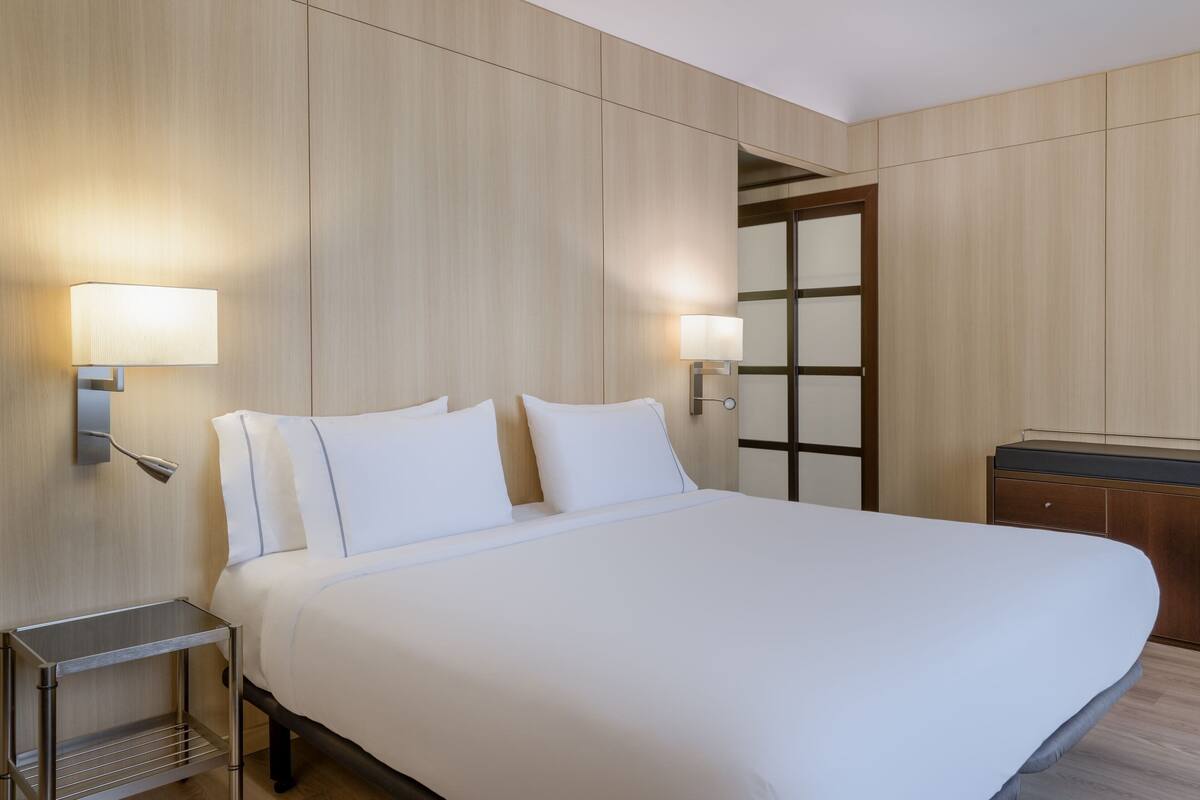 A hotel room with a large bed, two bedside lamps, a wooden wardrobe, and a small table.