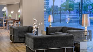 A modern lounge area with grey sofas, a coffee table, and a vase with white flowers. There are two lamps on the table, and a large window with a view of the street and trees. - AC Hotel Córdoba by Marriott (Córdoba)