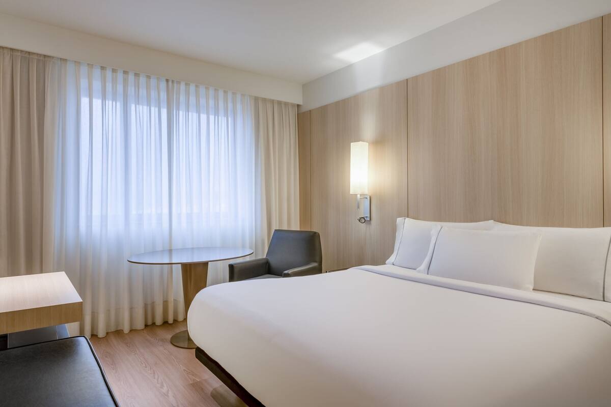A hotel room with a large bed, a small round table, a chair, and a wall-mounted lamp.