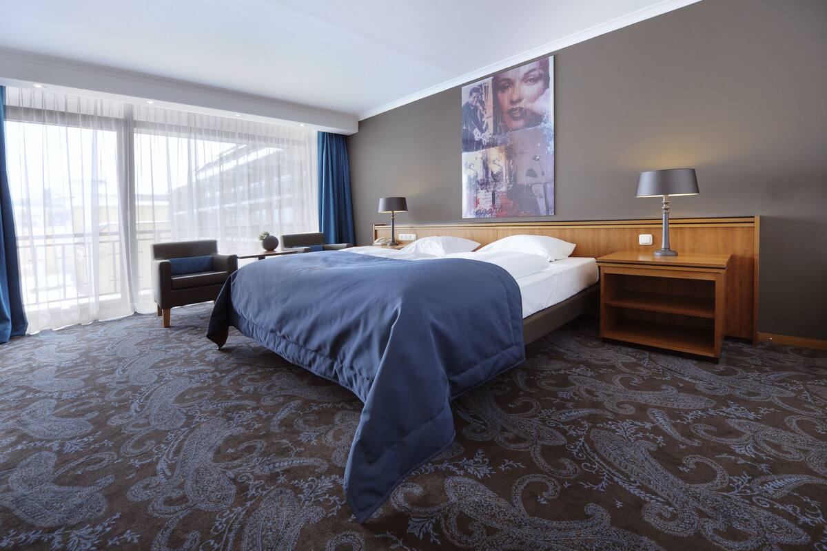 Habitación de hotel con una cama grande, dos mesitas de noche, una silla y un ventanal con cortinas.