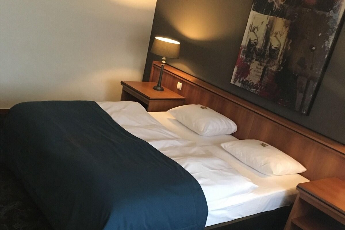 A hotel room with a bed, a nightstand with a lamp, a wooden headboard, and a patterned carpet.