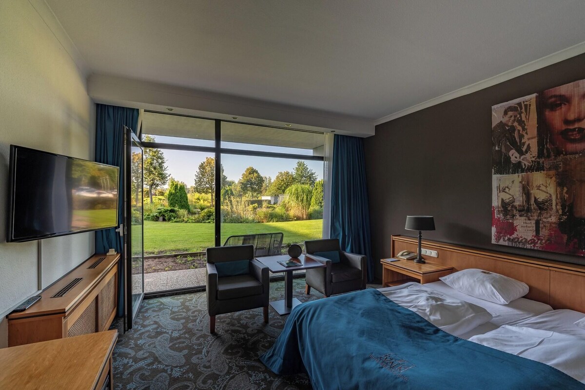 Ein Hotelzimmer mit Bett, Schreibtisch, Stuhl, Fernseher und einem großen Fenster mit Blick auf einen Garten.