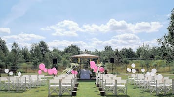 An outdoor wedding venue with white chairs arranged in rows, a gazebo in the center, and pink and white balloons.