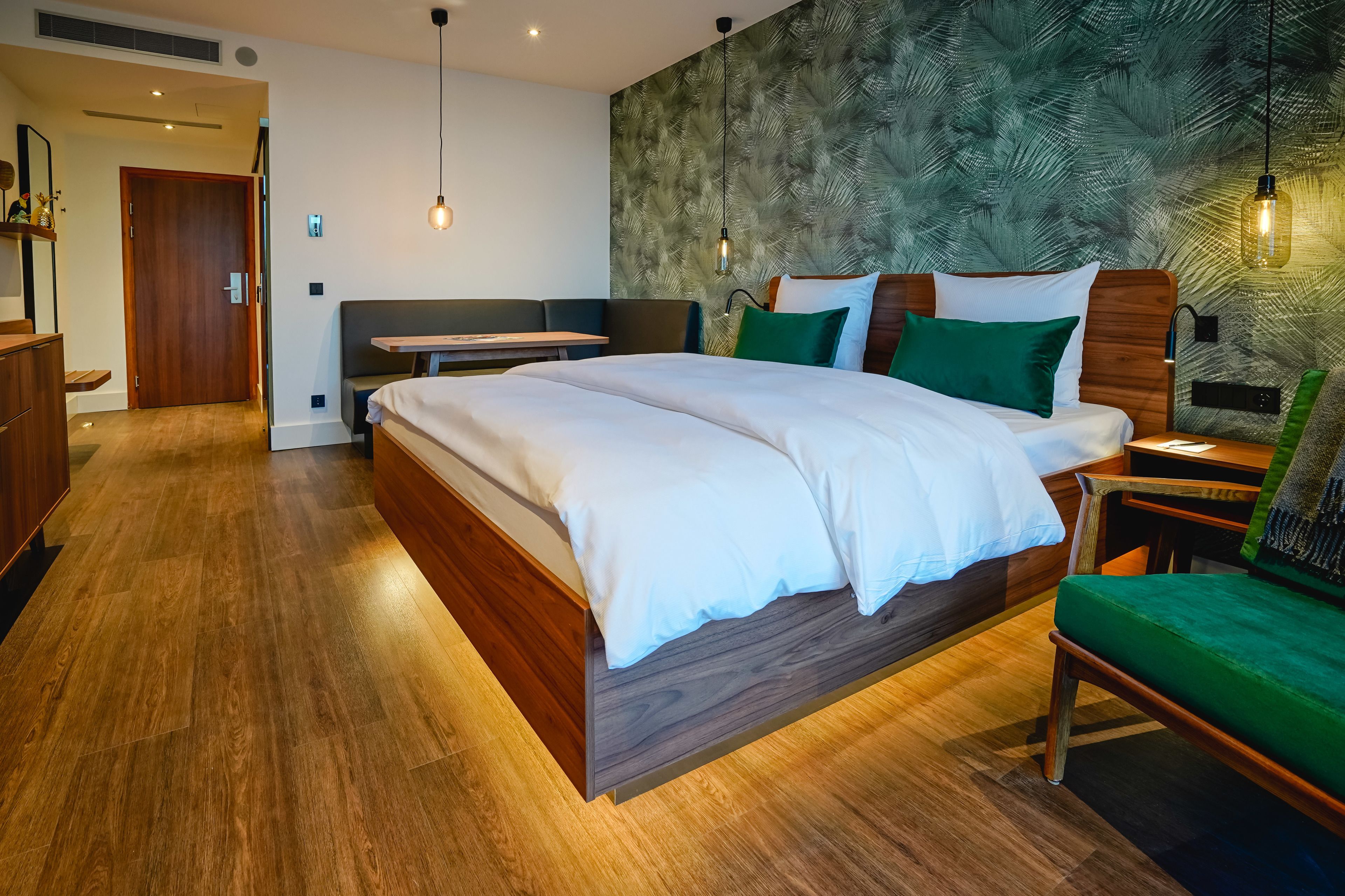 Una habitación de hotel moderna con una cama grande, una mesita de noche de madera, una silla verde y una pared con un diseño de hojas.