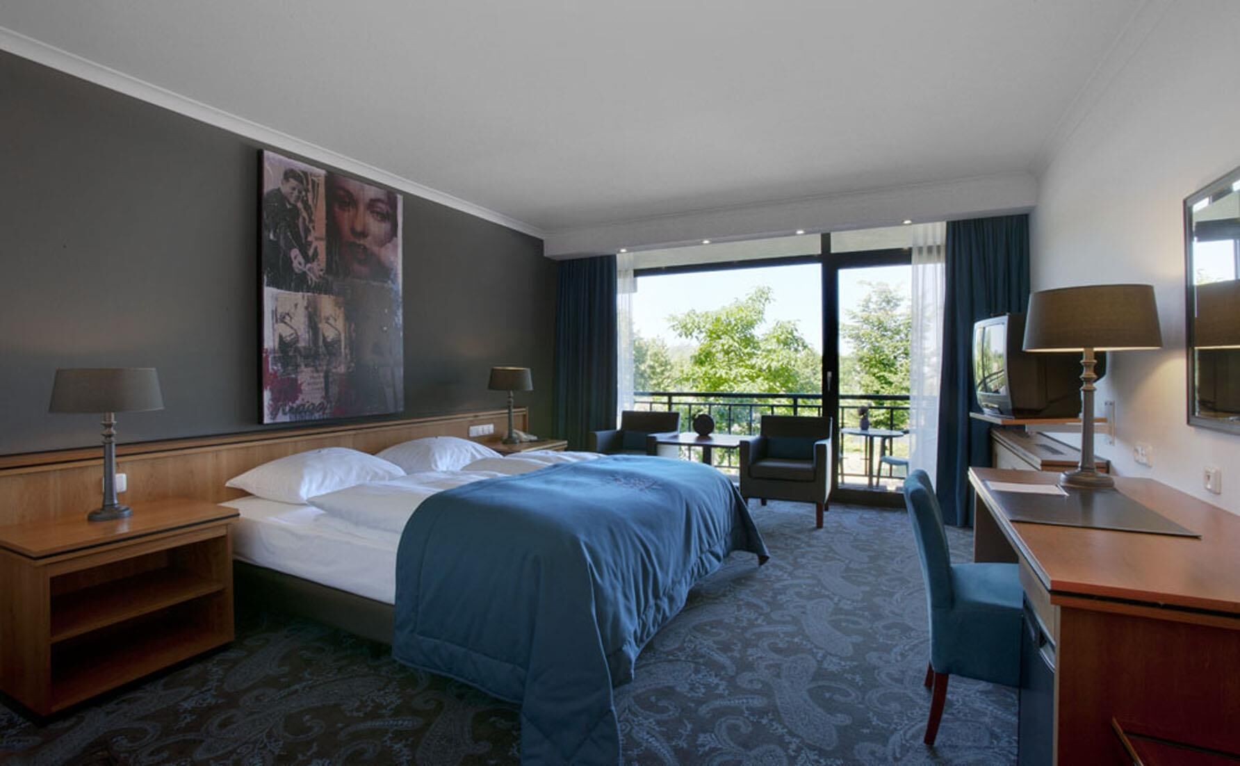 A hotel room with a bed, desk, chairs, and a balcony with a view.