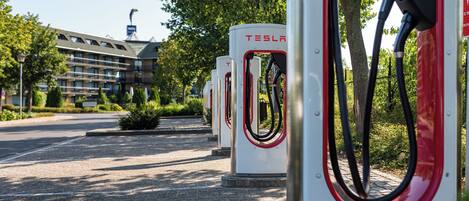 Una fila de estaciones de carga para vehículos eléctricos Tesla en un estacionamiento, con un hotel al fondo.