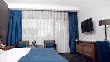 Una habitación de hotel con cama, dos sillas, una mesita, televisor y lámpara.
