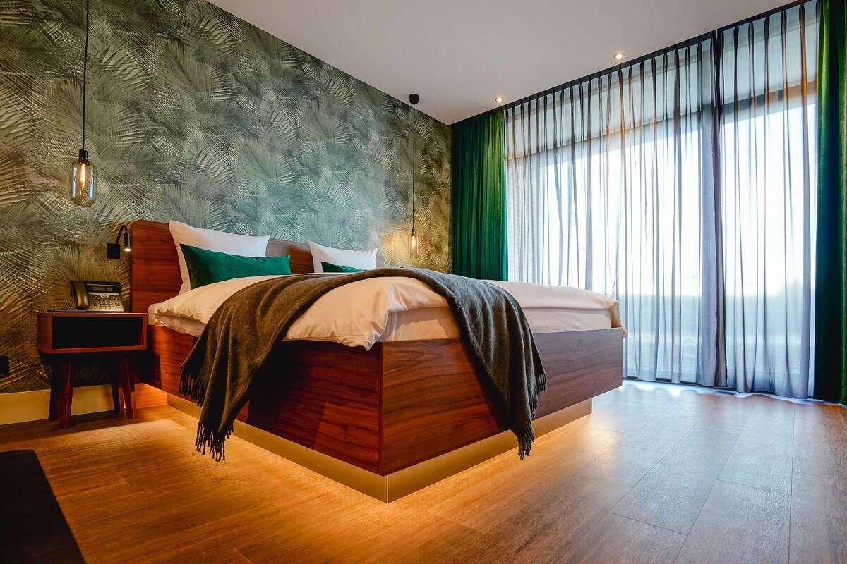 Un dormitorio moderno con una cama de madera, una mesita de noche, una lámpara y un amplio ventanal con cortinas.