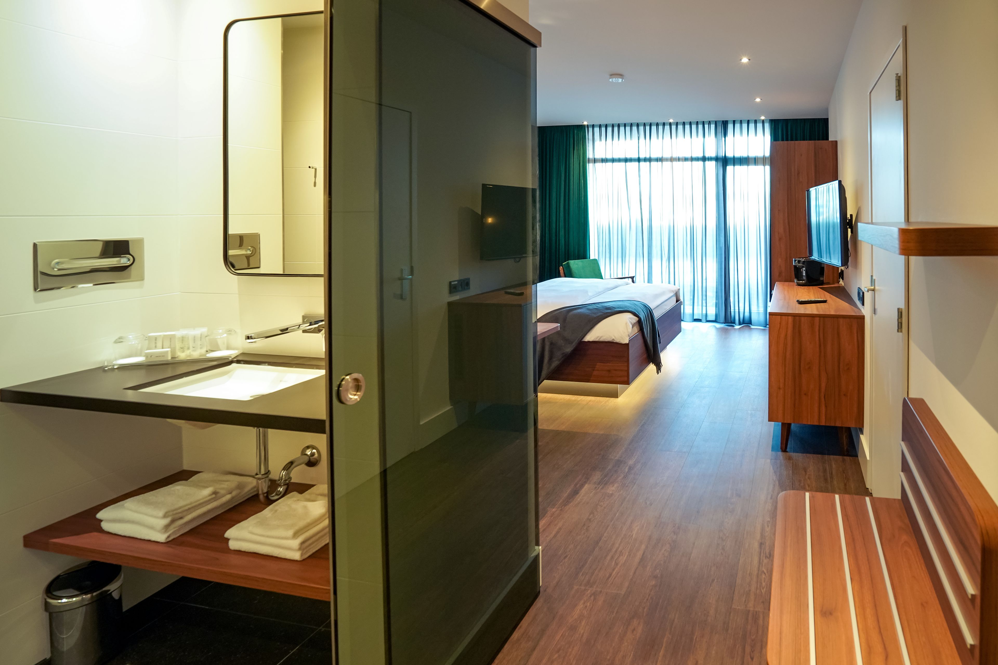 A modern hotel room with a glass-enclosed bathroom, a bed with a green headboard, a wooden desk, and a large window with curtains.