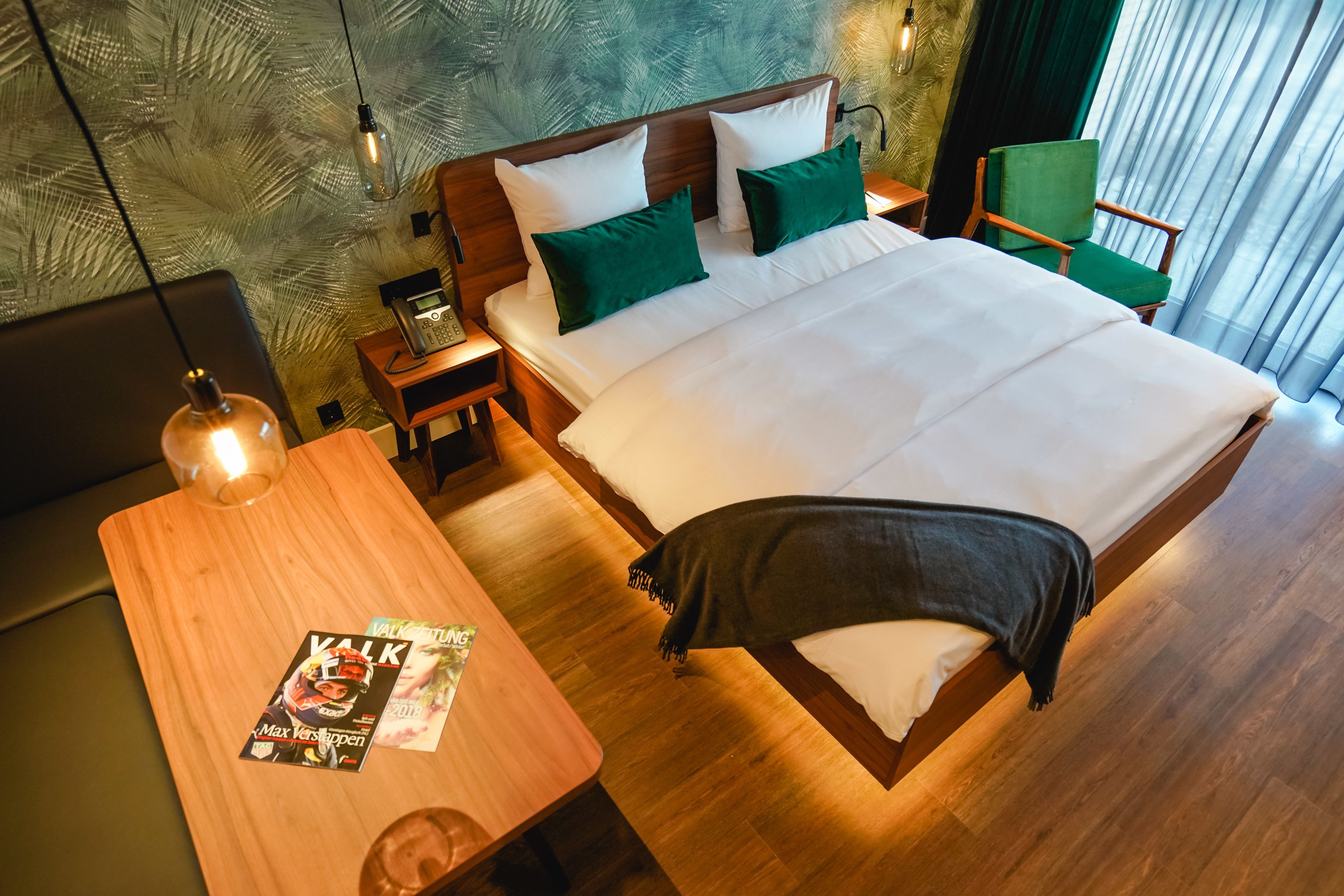 A hotel room with a wooden bed, green pillows, a wooden nightstand with a phone, a wooden table with a magazine, and a green chair.