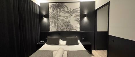 A modern hotel room with a large bed, a dark curtain, a wall-mounted artwork of palm trees, two bedside tables with lamps, and a large mirror.