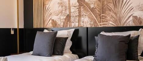 A hotel room with two beds, a large wall mural of tropical plants, a bedside table with a tray of toiletries, and a modern lamp.