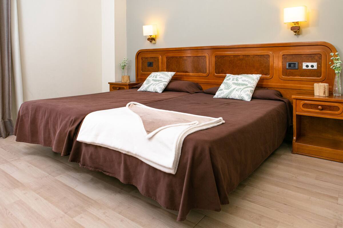 A neatly made bed with a brown bedspread and pillows, a bedside table with a lamp, and a nightstand with a vase.