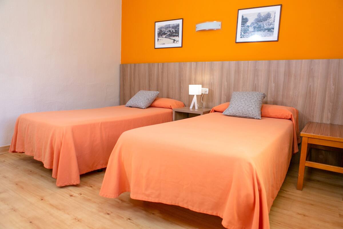 A room with two beds, each with an orange bedspread and matching pillows, a wooden nightstand with a lamp, and two framed pictures on the orange wall.