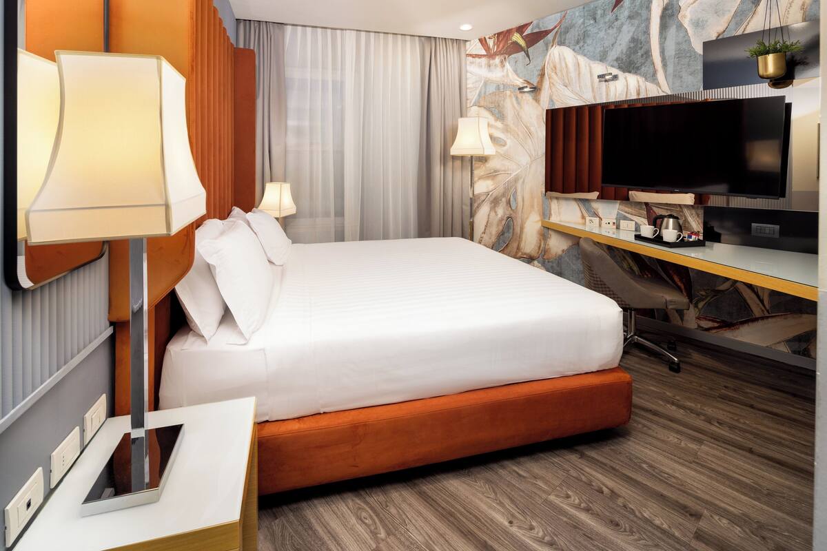 A modern hotel room with a large bed, a desk with a flat-screen TV, a lamp, and a small potted plant.