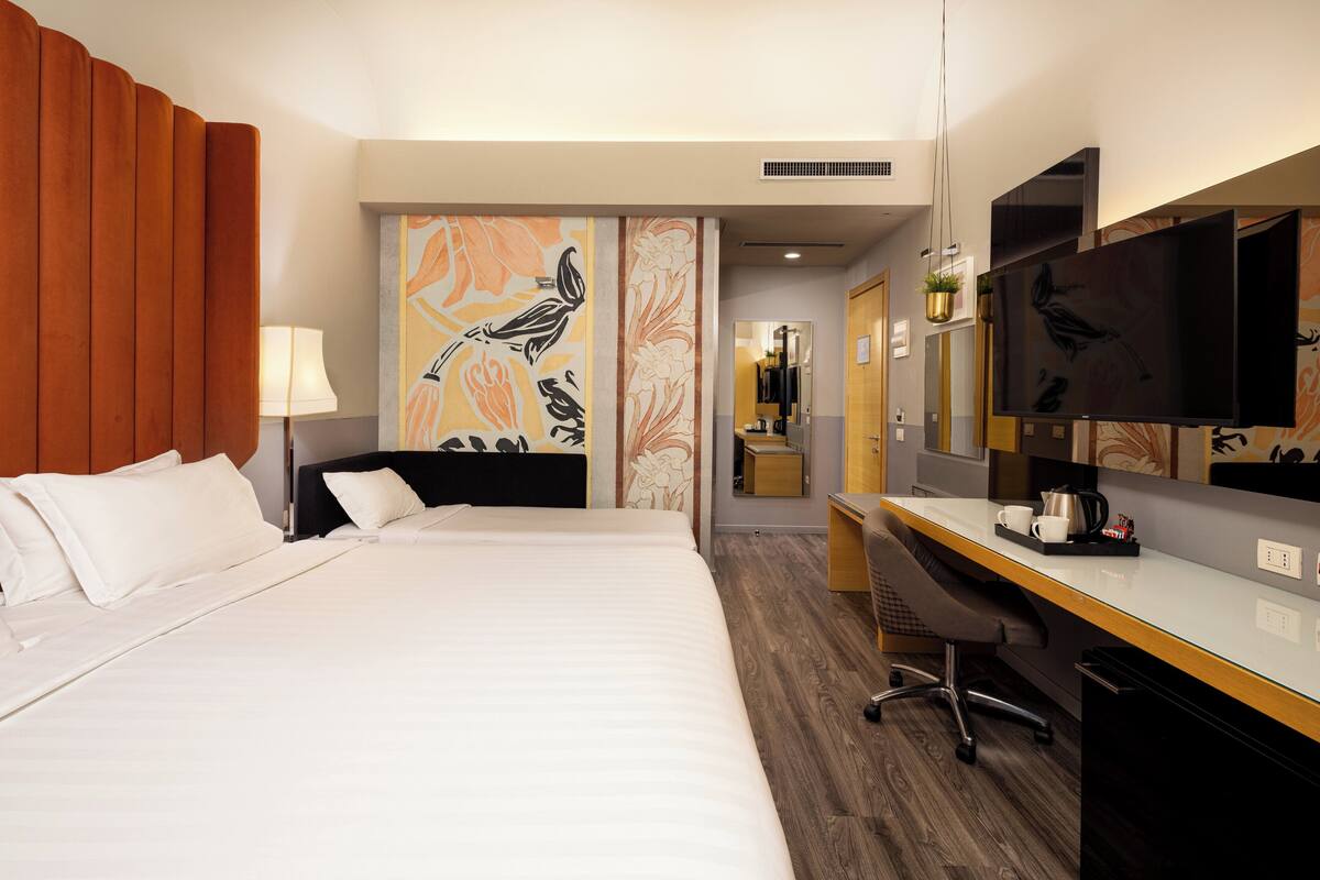 A hotel room with a bed, a desk, a chair, a TV, and a wall with a bird painting.