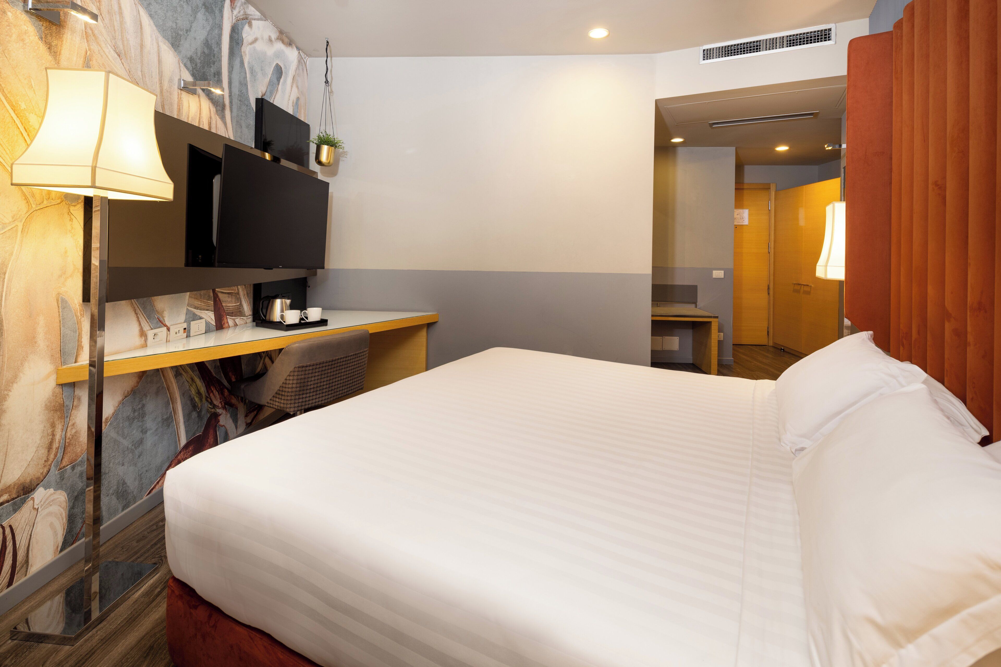 Habitación de hotel con cama, escritorio con lámpara, televisión de pantalla plana y planta en la pared.