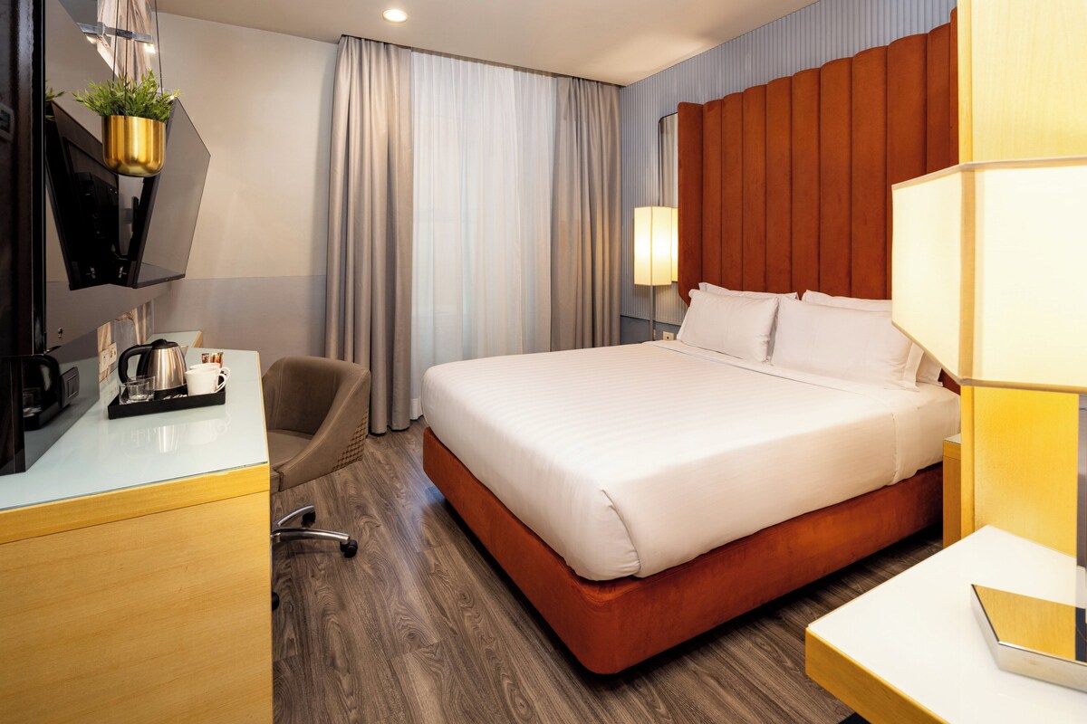 A hotel room with a bed, a desk with a coffee maker, a chair, a TV, and a plant.