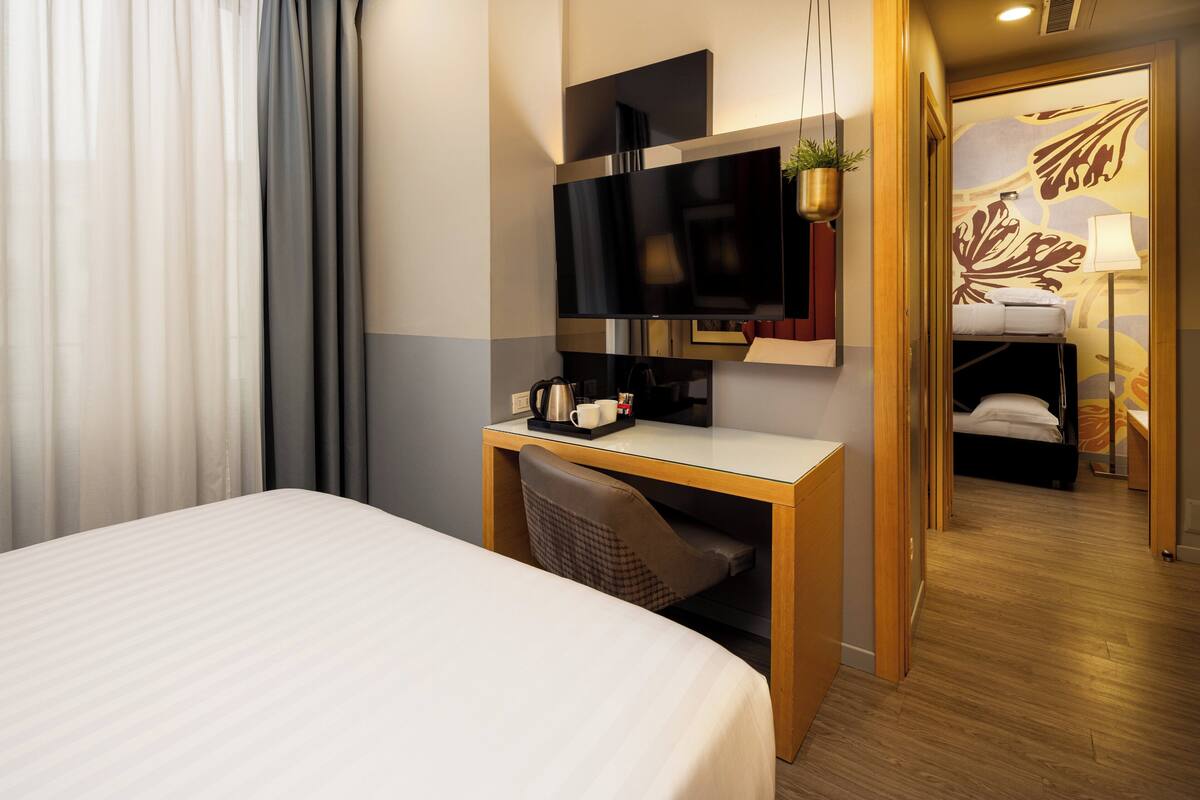 Quarto de hotel com cama, escrivaninha com televisão, cafeteira e planta.