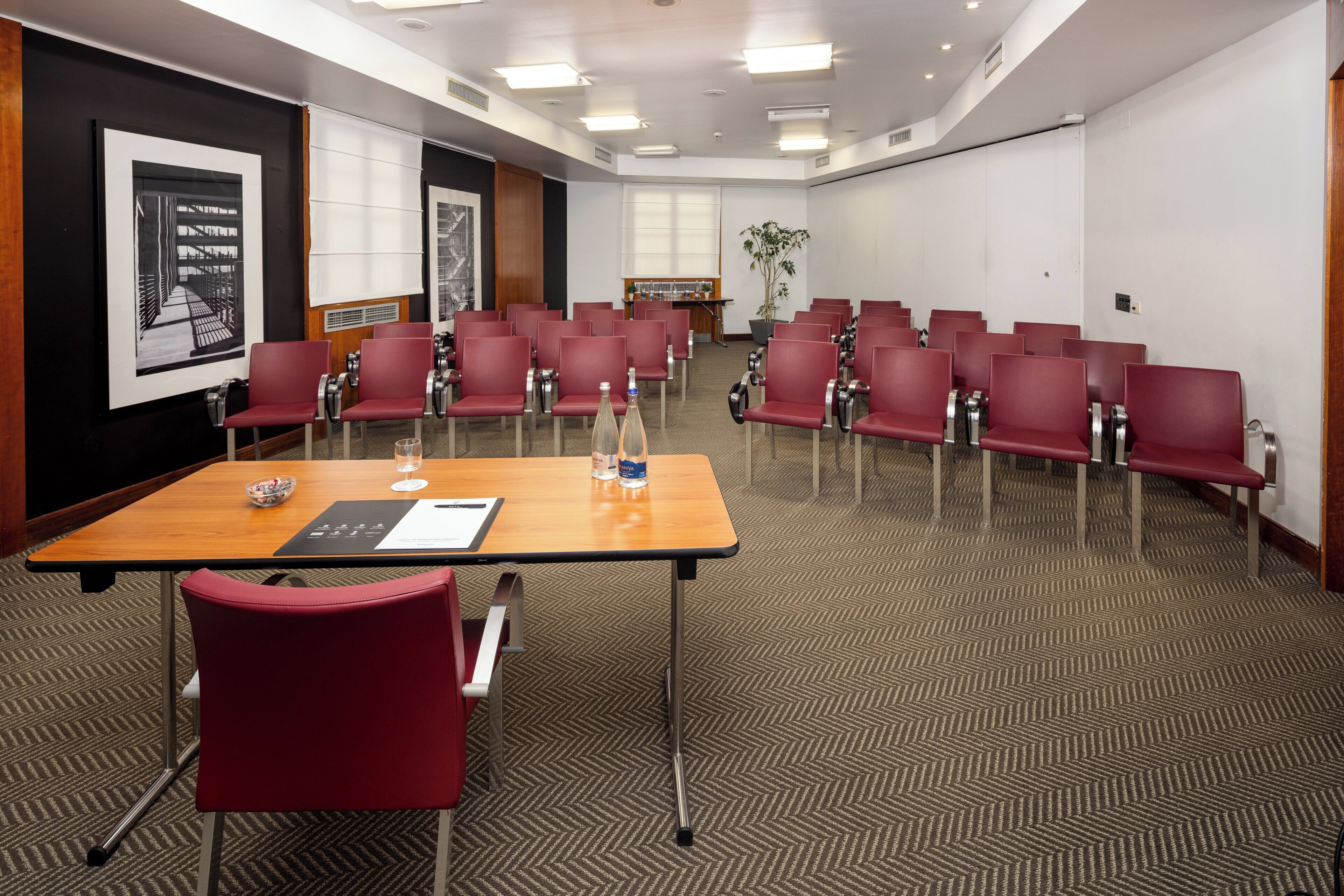 A conference room with a long table, red chairs, a projector screen, and a plant.